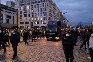 Eine Gruppe von Menschen steht vor einem Lastwagen auf einer Straße, umgeben von Gebäuden, Laternenmästen, Bäumen und einem wolkenverhangenen Himmel, einige tragen Mötzen und Masken, und ein Band mit einem Pfahl im Vordergrund.