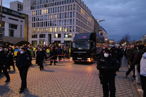Eine Gruppe von Menschen steht vor einem Lastwagen auf einer Straße, umgeben von Gebäuden, Laternenmästen, Bäumen und einem wolkenverhangenen Himmel, einige tragen Mötzen und Masken, und ein Band mit einem Pfahl im Vordergrund.