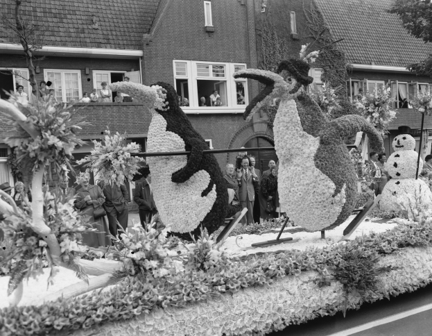 Schwarzes und weißes Foto eines Paradewagens, der mit Blumen und Pinguinen geschmückt ist, mit Menschen, Bäumen und Gebäuden im Hintergrund.