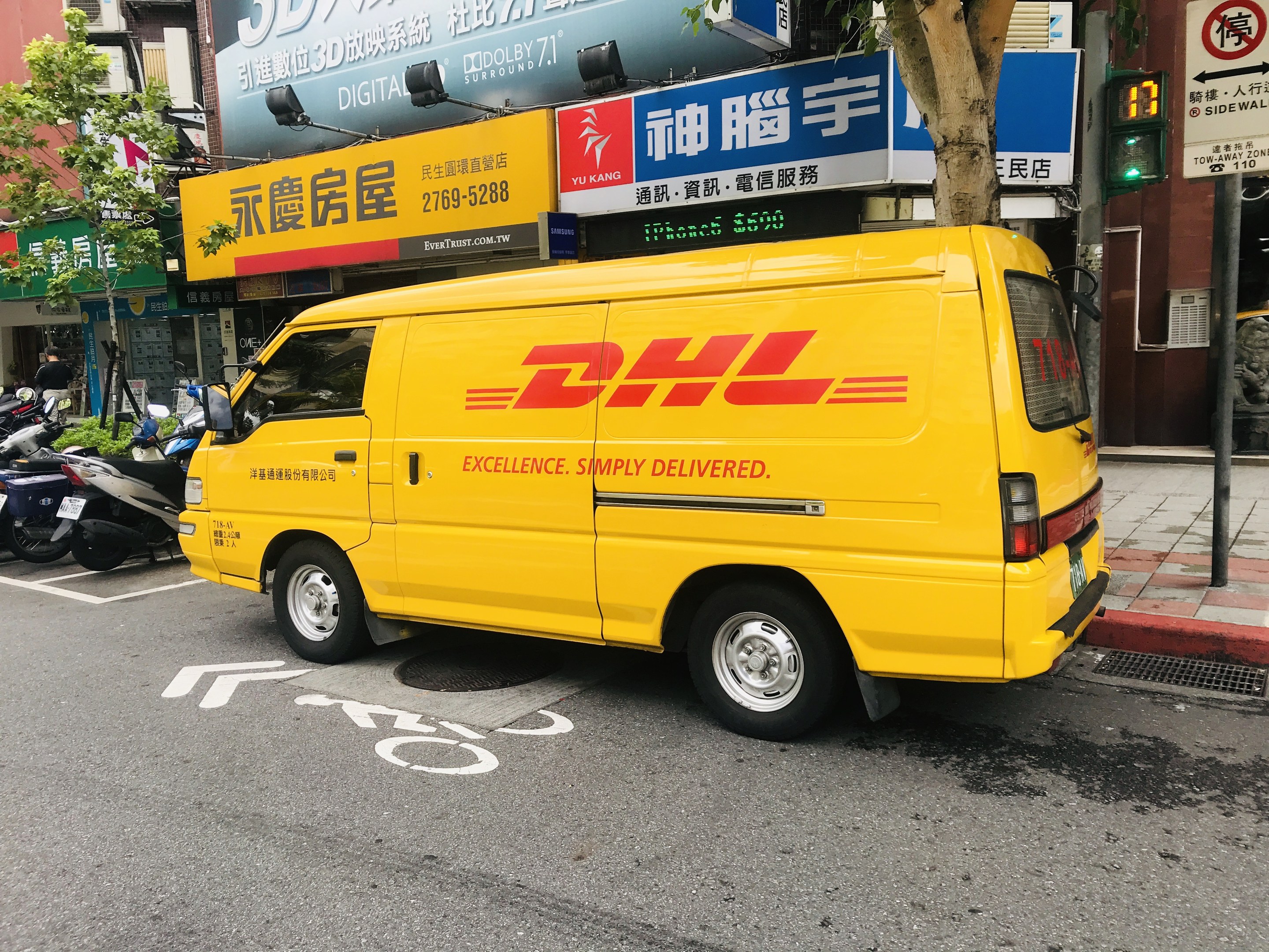 Gelbes DHL-Lieferfahrzeug auf der Straßenseite geparkt, umgeben von Gebäuden, Bäumen, Schildern, Mästängen, Ampeln und einem Füßgänger auf dem Gehweg.