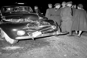 Eine Schwarz-Weiß-Szene eines beschädigten Autos am Straßenrand mit einer Gruppe besorgter Menschen drumherum.