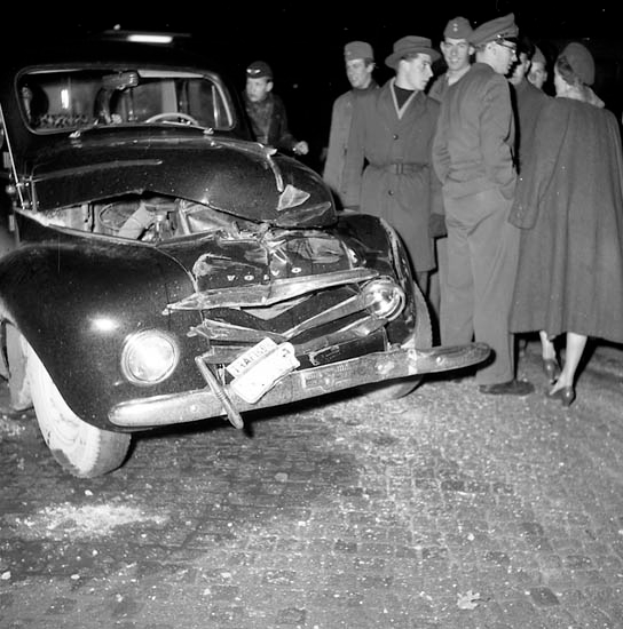 Eine Schwarz-Weiß-Szene eines beschädigten Autos am Straßenrand mit einer Gruppe besorgter Menschen drumherum.