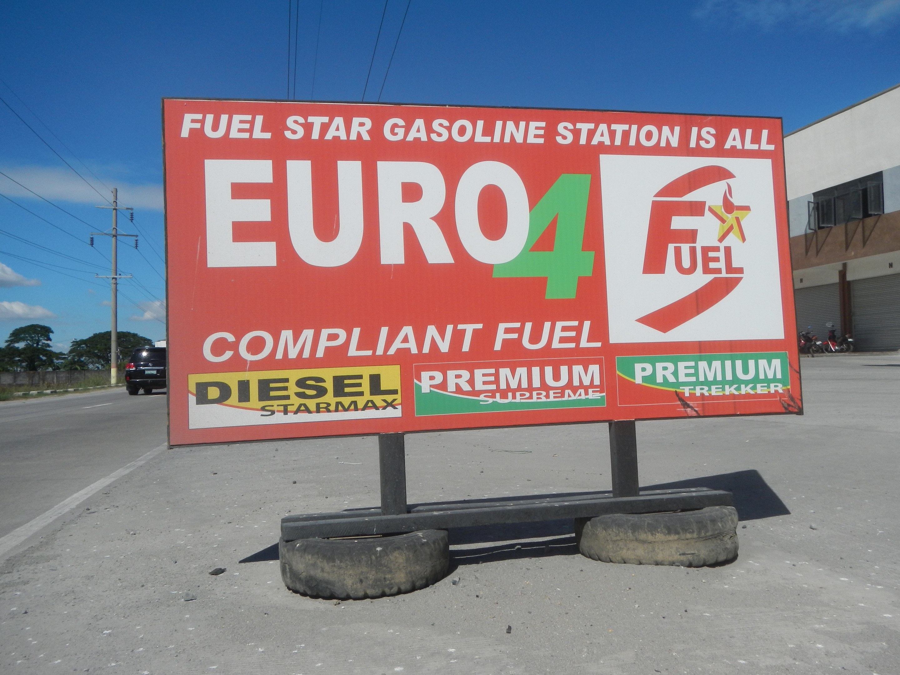 Eine Tankstelle mit einem "Euro4 Kraftstoff"-Schild im Vordergrund, Fahrzeuge auf der Straße, Strommasten mit Drähten, Bäume, ein Gebäude auf der rechten Seite und ein Himmel oben.