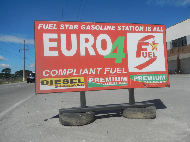 Eine Tankstelle mit einem "Euro4 Kraftstoff"-Schild im Vordergrund, Fahrzeuge auf der Straße, Strommasten mit Drähten, Bäume, ein Gebäude auf der rechten Seite und ein Himmel oben.