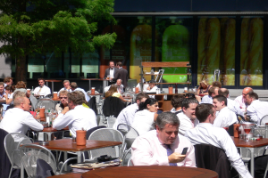 Eine Kantine mit Gruppen von Menschen an Tischen sitzend und sich unterhaltend, zwei Individuals in schwarzen Anzügen in der Mitte stehend, ein Flaschencontainer rechts, ein Baum links und Saftgläser und Flaschen auf den Tischen.