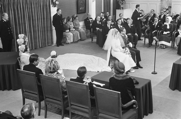Schwarzes und weißes Foto einer Hochzeitszeremonie mit sitzenden und stehenden Gästen, einem Mann, der in ein Mikrofon spricht, einer Frau mit einem Blumenstrauß, einem Podium mit einem Mikrofon auf der rechten Seite, Blumenschmuck und einem Vorhang auf der linken Seite, einem gerahmten Foto an der Wand und einer Tür im Hintergrund.