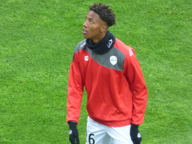 Ein junger Mann in einer roten Jacke und weißen Shorts steht auf einem Fußballfeld und trägt Baseballhandschuhe, wobei er entschlossen nach rechts schaut.