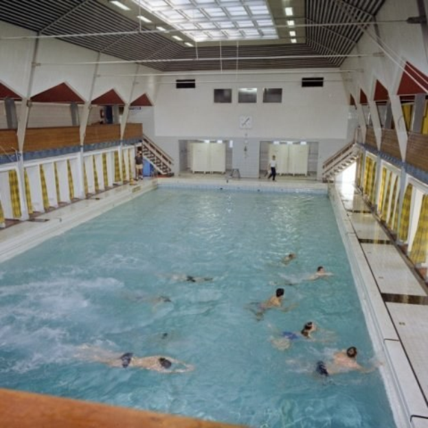 Großes Hallenbad mit schwimmenden Menschen, flankiert von Säulen und mit einer Uhr an der Wand sowie heller Deckenbeleuchtung.