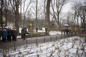 Gruppe von Menschen in militärischer Kleidung, die vor einem Denkmal mit einer Tafel und Grünzeug im Vordergrund, Bäumen und Himmel im Hintergrund stehen.