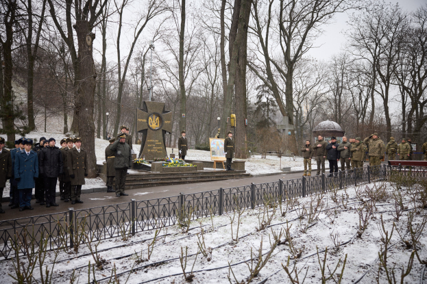 Gruppe von Menschen in militärischer Kleidung, die vor einem Denkmal mit einer Tafel und Grünzeug im Vordergrund, Bäumen und Himmel im Hintergrund stehen.