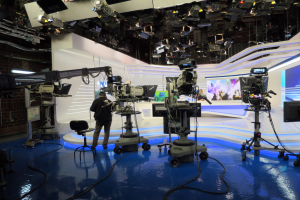 Ein Mann steht vor einem Fernseher und hält ein Mikrofon in der Hand, umgeben von Kameras, einem Stuhl, einem Bildschirm, einer Wand und einer Decke mit Lampen, was auf eine Live-Übertragung hindeutet.