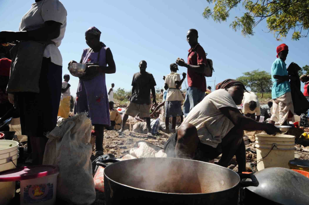 Eine Gruppe von Menschen bereitet Essen im Freien in einer ländlichen Gegend zu, mit einem großen Topf, Eimern und anderen Gegenständen auf dem Boden unter einem klaren blauen Himmel.