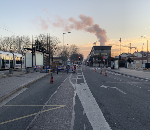 Straßenszene mit Verkehrskegeln, einem Bus, Laternenpfählen, Bäumen, Gebäuden, Kränen und Rauch, der aus einem entfernten Gebäude aufsteigt.