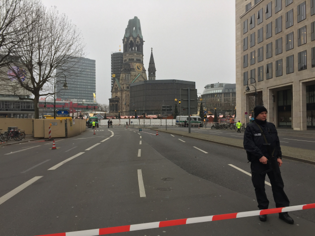 Ein Polizist in schwarzer Uniform und Mütze steht in einer Berliner Straße umgeben von Verkehrskegeln, Fahrrädern, Laternen, Bäumen und Gebäuden, mit einem Uhrenturm und einem klaren blauen Himmel im Hintergrund.
