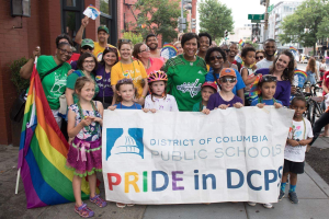 Gruppe von Menschen, die draußen vor einem Banner mit der Aufschrift "District of Columbia Public Schools Pride in DCPS" stehen, mit Kindern, die eine Fahne halten, einem Radfahrer, Bäumen, Gebäuden, Straßenschildern, Ampeln und einem bewölkten Himmel.