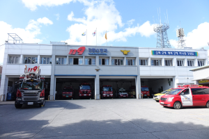 Feuerwache mit mehreren Feuerwehrautos vor dem Gebäude geparkt, ein Gebäude mit Fenstern und Text, Flaggen mit Stangen im Hintergrund, bewölkter Himmel und eine ferne Turm.