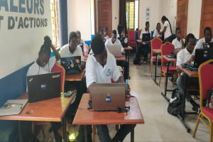 Eine Gruppe von Menschen sitzt an Tischen mit Laptops in einem Klassenzimmer, mit Taschen auf dem Boden und Text an der linken Wand, was auf einen Online-Kurs in Ghana hinweist.