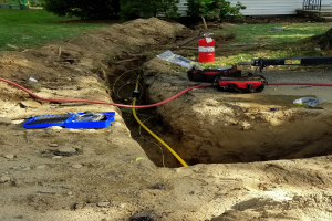 Großes Loch im Boden mit angeschlossenem Schlauch, umgeben von Werkzeugen einschließlich eines Feuerlöschers und Rohren, in einem Wohngebiet mit Bäumen, Pflanzen, Gras und einem Haus.