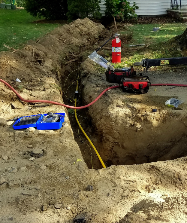 Großes Loch im Boden mit angeschlossenem Schlauch, umgeben von Werkzeugen einschließlich eines Feuerlöschers und Rohren, in einem Wohngebiet mit Bäumen, Pflanzen, Gras und einem Haus.