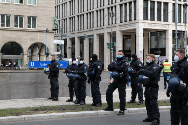 Gruppe von Polizisten in schwarzen Uniformen und Masken vor einem Berliner Gebäude mit Glasfenstern und Säulen, einige halten Helme, mit Laternenmasten, Verkehrsampeln, Texttafeln, einer Statue und anderen Menschen im Hintergrund auf einem grasbewachsenen Boden.