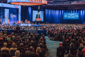 Eine große Menschenmenge sitzt auf Stühlen, die einer Bühne zugewandt sind, mit ein paar Menschen, die darauf stehen, blauen Vorhängen, einem Bildschirm und einem orangefarbenen Schild oben, das die CPAC-Veranstaltung am 25. Oktober anzeigt.