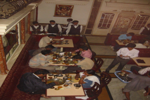 Eine Gruppe von Menschen an Tischen essend, mit einigen im Hintergrund bedienend, ein Foto mit Blumen auf der Wand und eine Tür im Hintergrund.