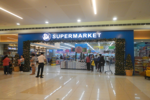 Eine Gruppe von Menschen steht vor einem Supermarkt, einige halten Taschen, mit einem Einkaufswagen, einem Topf mit einer Pflanze, Regalen mit Gegenständen, Schildern und einer Decke mit Beleuchtung.