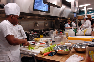 Eine Gruppe von Köchen in einer Küche, mit einem im Vordergrund in einer weißen Schürze und Mütze, der ein Messer hält und von verschiedenen Utensilien, Schüsseln, Gemüse, einem Handy, Flaschen und anderen Gegenständen umgeben ist, während im Hintergrund ein Fernseher und Deckenleuchten zu sehen sind.