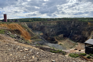 Großer Tagebau in einer Schotterfläche umgeben von Bäumen, Gebäuden und einem Schuppen unter einem bewölkten Himmel mit Steinen und Pflanzen auf dem Boden.
