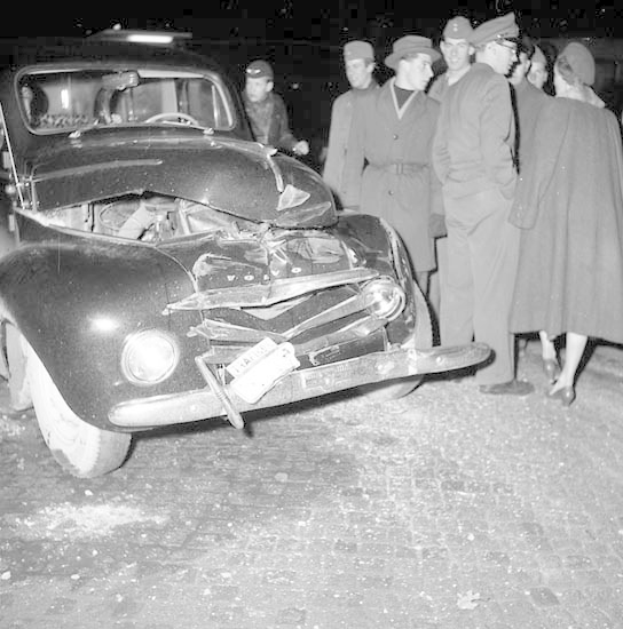 Schwarz-weißes Bild eines schwer beschädigten Autos am Straßenrand mit eingedellter Motorhaube, zersplitterten Fenstern und einer Gruppe von Menschen drumherum, die besorgt aussehen.