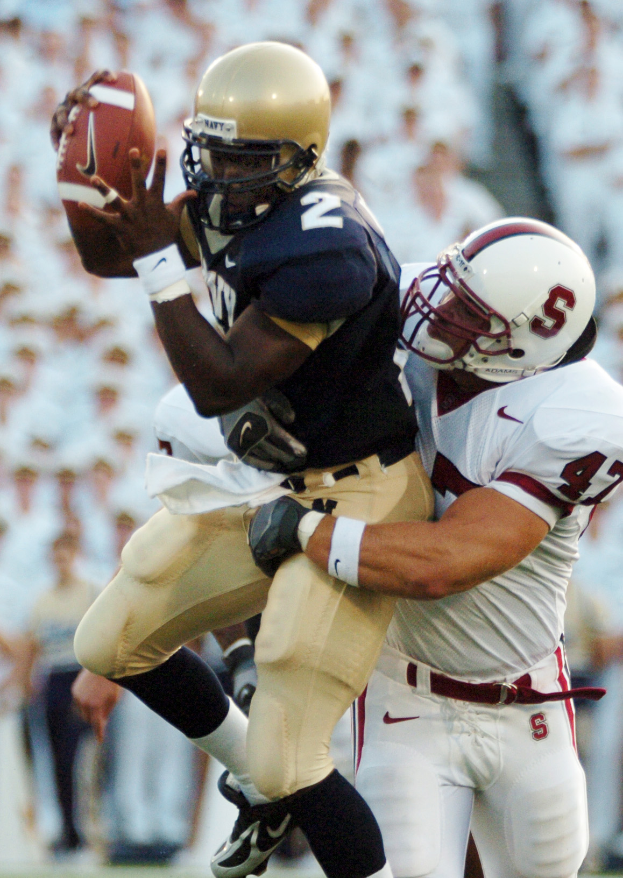 Ein Fußballspieler fängt einen Ball in der Luft, während er von einem anderen Spieler getackelt wird, beide tragen Sportkleidung und Helme, mit einem unscharfen Hintergrund.