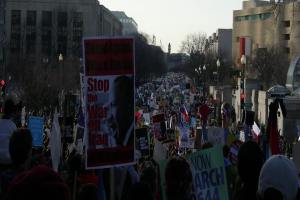 Eine Gruppe von Menschen, die auf einer Straße protestieren und Plakate halten, mit Gebäuden, Bäumen, Laternenmasten und dem Himmel im Hintergrund.