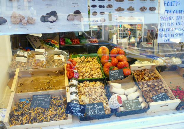 Marktstand-Vitrine mit Tabletts voller Gemüse, Flaschen und anderen Lebensmitteln, Papieren mit Text auf der Scheibe und Menschen im Hintergrund.