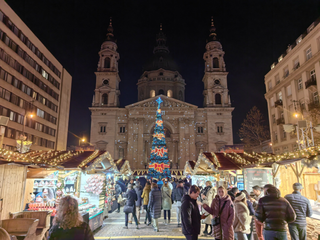 Ein belebter Weihnachtsmarkt mit festlichen Ständen und Lichtern vor einer Kirche bei Nacht, umgeben von Gebäuden, Bäumen und einem sternenklaren Himmel.