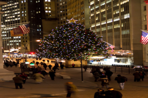 Ein Weihnachtsbaum auf einer Straße mit Menschen darauf, flankiert von zwei Fahnenmasten mit Fahnen und beleuchteten Gebäuden im Hintergrund.