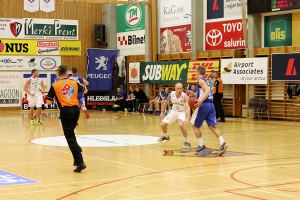 Ein Basketballfeld mit Spielern im Vordergrund und einer hölzernen Wand mit Plakaten und Bannern im Hintergrund.