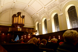 The interior of a church with men and women seated, candles in front of them, and white walls and roof.