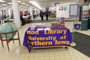 Eine Expo-Situation mit einem Tisch, auf dem verschiedene Landesflaggen, ein Karton, Papiere und ein Banner liegen, eine Weltkugel links daneben, ein Anschlagbrett rechts daneben mit ein paar Menschen daneben und mehreren Tischen und Stühlen in der Ferne.