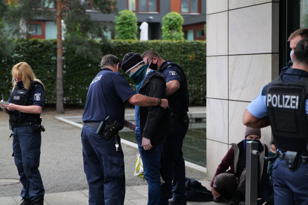 Gruppe von Polizisten vor einem Gebäude stehend, mit einer Frau, die ein Buch und einen Stift hält, umgeben von sitzenden und stehenden Personen, mit Bäumen und Gebäuden im Hintergrund.