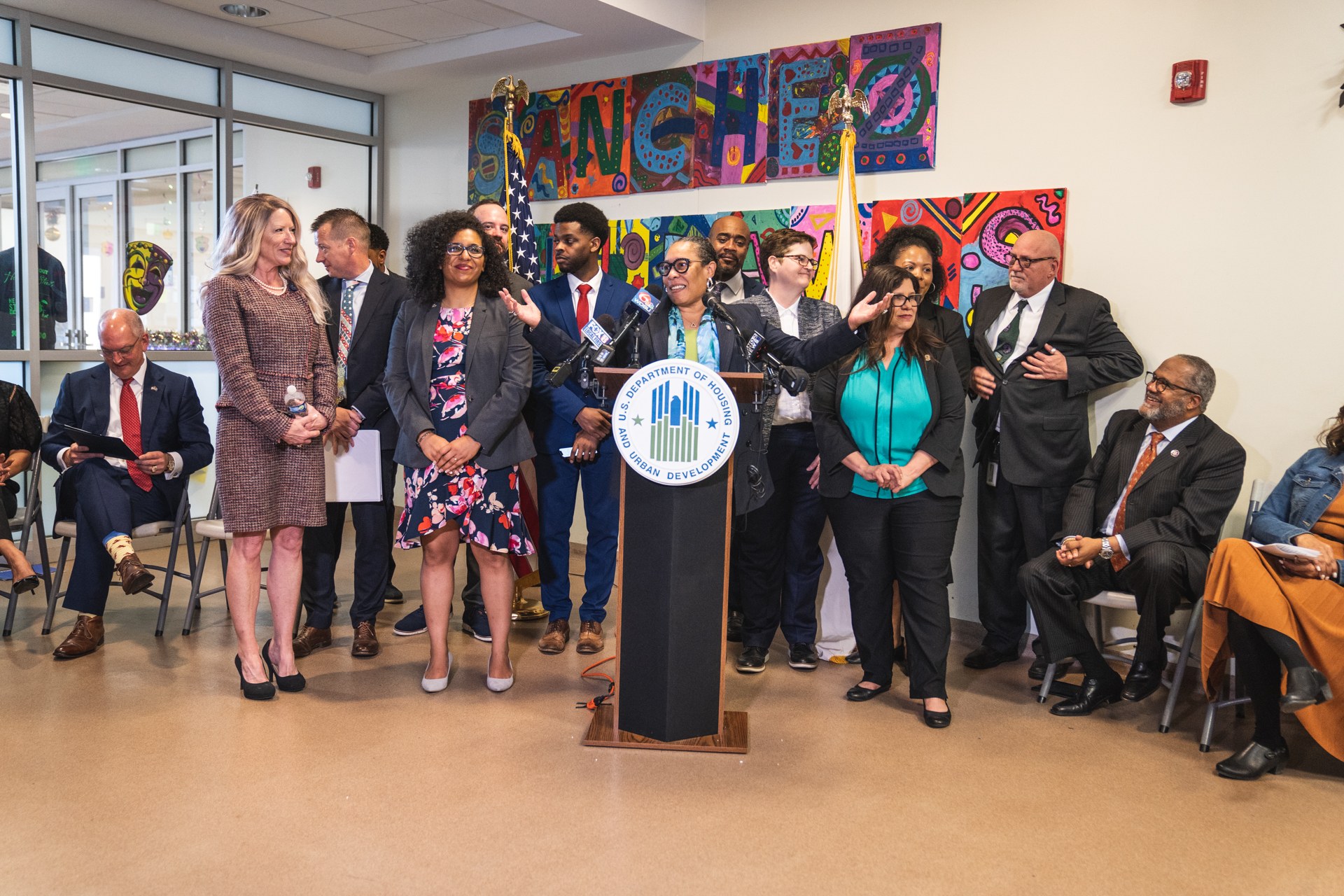 Gruppe von Menschen vor einem Rednerpult mit einem Mikrofon und einer Tafel mit der Aufschrift "Sancho-Stadtrat tritt dem Stadtrat bei", mit sitzenden und stehenden Teilnehmern, Flaggen, einem Feuerlöscher und sichtbaren Deckenleuchten.