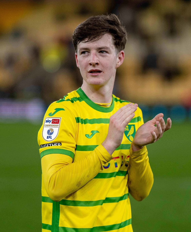 Ein junger Mann in einem gelb-grünen T-Shirt steht auf einem Fußballfeld und klatscht in die Hände, mit einem unscharfen Hintergrund.