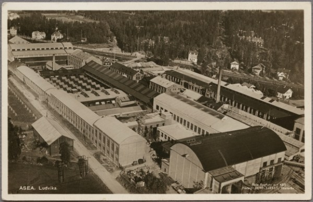 Schwarzes und weißes Foto einer Industrieanlage mit Gebäuden, Schuppen, Bäumen, Masten, Straßen und anderen Gegenständen mit Text unten.