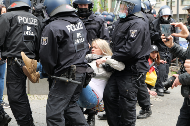 Polizeibeamte in Einsatzausrüstung begleiten eine Frau in einem weißen und blauen Kleid, während jemand das Geschehen fotografiert.