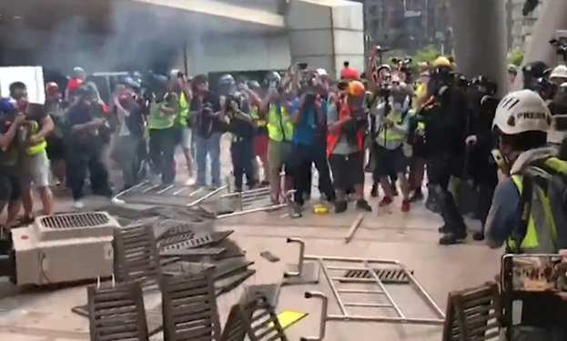 Menschen in Helmen und Jacken stehen vor einem rauchenden Gebäude während einer Stadtprotest, einige halten Kameras, während Trümmer den Boden bedecken.