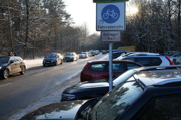 Verkehrsstau auf einer verschneiten Straße mit parkenden Autos, ein Verkehrsschild im Vordergrund, Tannen und einen klaren Himmel im Hintergrund.