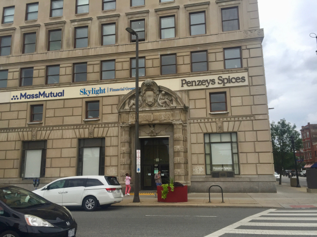 Großes Gebäude mit parkenden Autos davor, Straßenlaternen, Verkehr, Fußgänger auf dem Gehweg, Grünfläche und bewölktem Himmel, identifiziert als Hauptsitz der Massachusetts Mutual Financial Group.