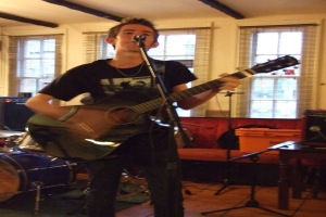 Ein Mann in schwarzem T-Shirt und Hose steht mit einer Gitarre und einem Mikrofon auf einem Ständer, mit einer blauen Trommel in der Ecke, einer Couch mit Gegenständen darauf und durchsichtigen Vorhängen im Hintergrund unter einer Decke.