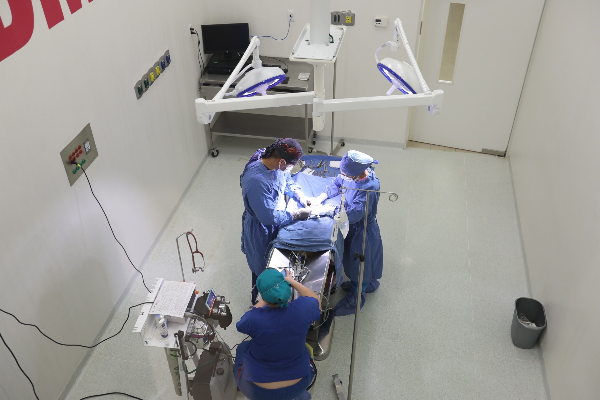 Chirurgen führen einen Eingriff an einem Patienten in einem Operationssaal durch, mit medizinischer Ausrüstung an einer Wand links und einem Objekt auf dem Boden rechts.