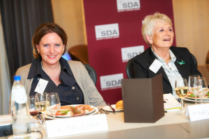 Zwei Frauen sitzen an einem Tisch mit Essen, Getränken und diversen Gegenständen, sie lächeln, mit einem Banner, Stühlen und Vorhängen im Hintergrund.