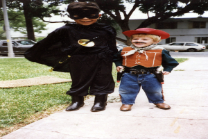 Kinder in verschiedenen Kostümen stehen auf einer Straße mit Fahrzeugen, Gebäuden, Bäumen und Straßenlaternen im Hintergrund.